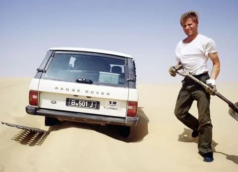 Amadeus Matzker mit Schaufel neben einem Range Rover im Wüstensand.